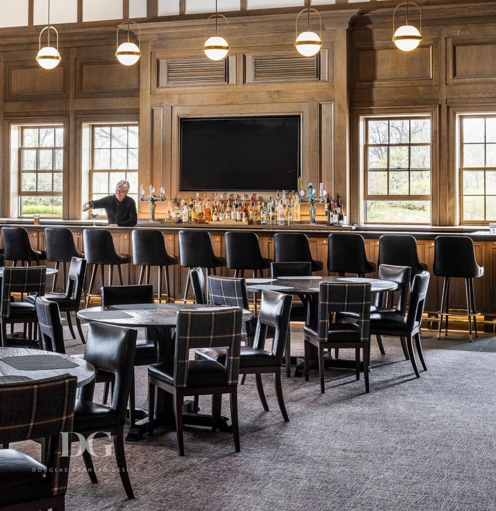 Looking across wood panelled dining room at a bar with leather bar stools, globe pendant lights and a barman pouring a drink