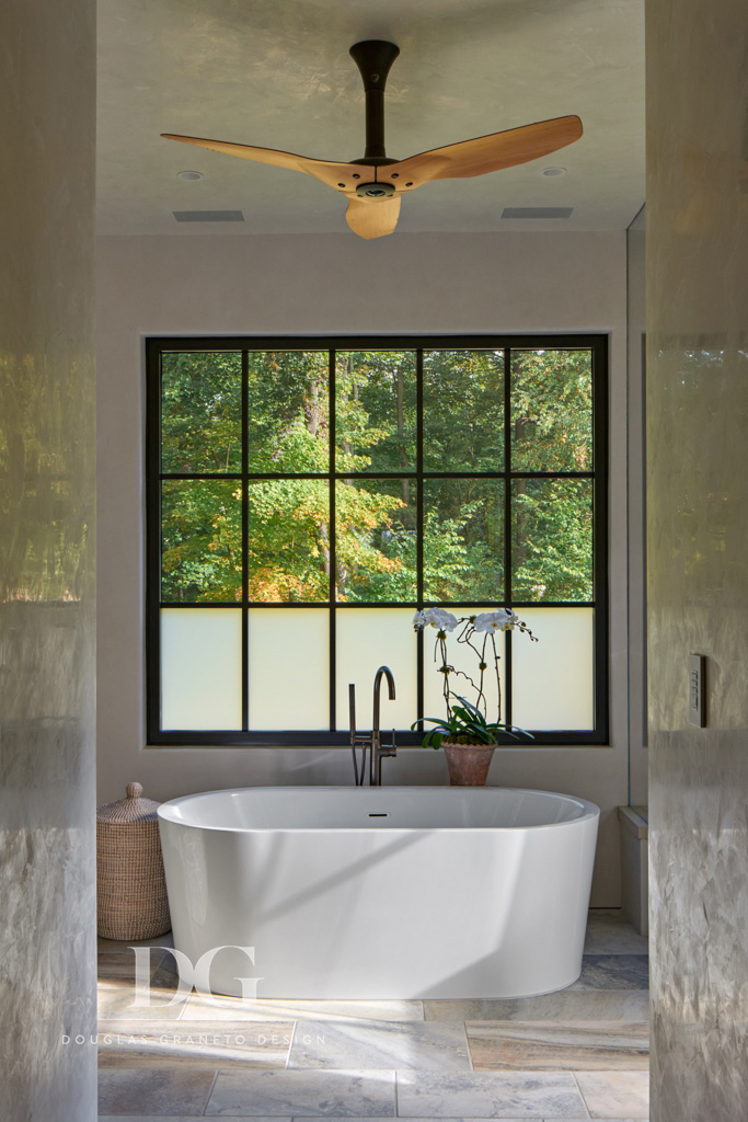 A modern white bathtub in front of a large black trimmed window, with stone flooring and a Bigass Fans ceiling fan