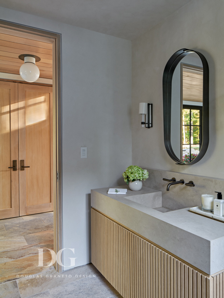 Side view of a custom vanity with pale wood beadboard millwork by Rock Ridge Fine Cabinetry, stone top and built-in sink, oval mirror by Swiss Welding, Urban Electric light fixtures