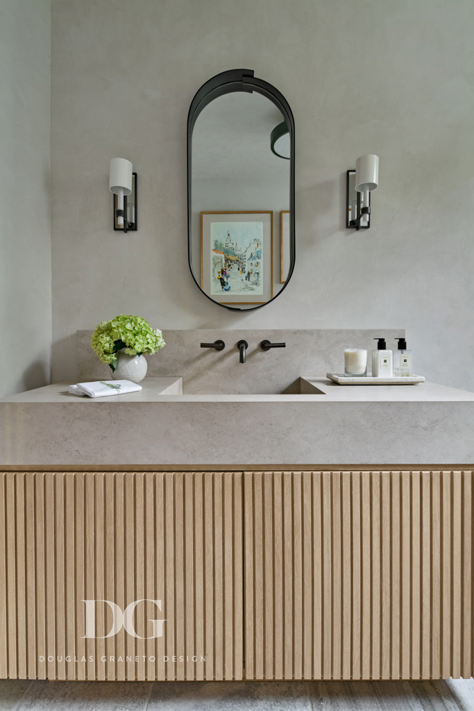 Front view of a custom vanity with pale wood beadboard millwork by Rock Ridge Fine Cabinetry, stone top and built-in sink, oval mirror by Swiss Welding, Urban Electric light fixtures