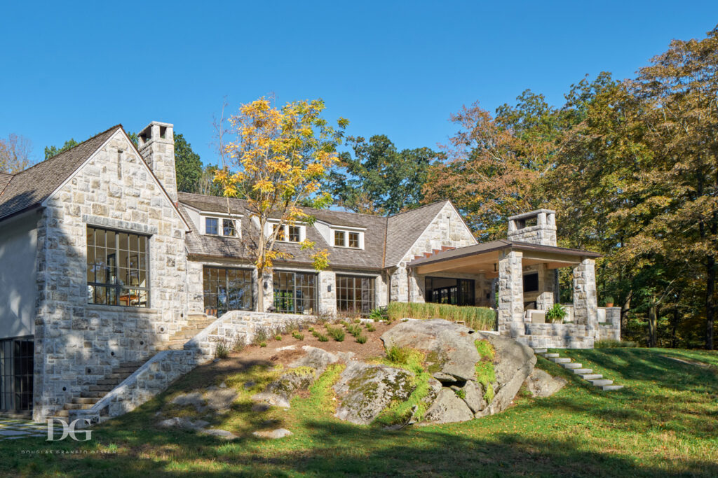 A large stone clad house set amongst grass and trees with a large stone outcrop in front