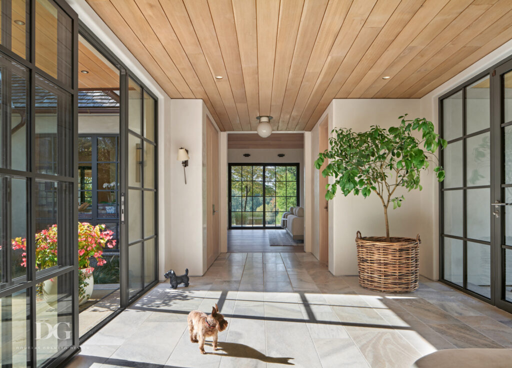 A large home entryway with stone floor, black trimmed sliding glass doors to patio and a tree in a large basket. Urban Electric light fixtures.