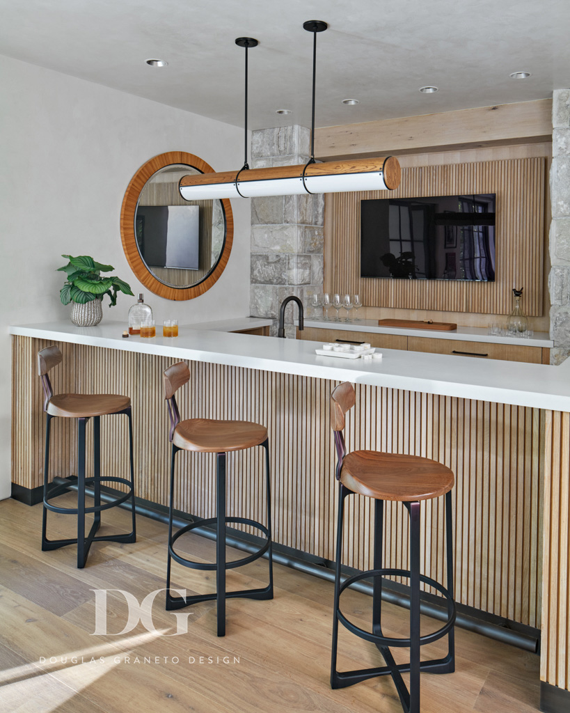 A custom home bar with white counter and custom pale wood millwork. Holly Hunt counter stools and Rock and Hill light fixture