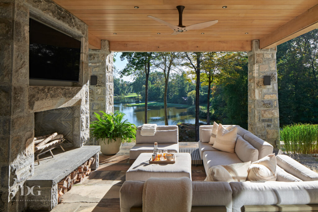 Large covered patio with stone clad fireplace, wood paneled ceiling with fan and cream sectional with Grayson De Vere pillows and throws