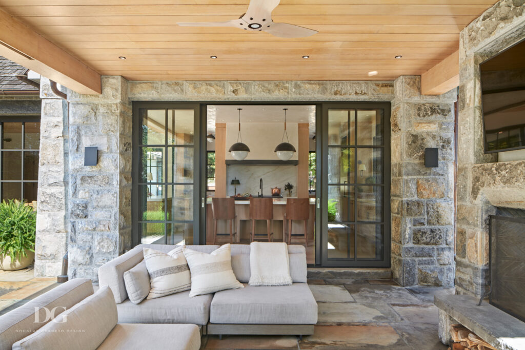 View from covered patio looking through sliding glass doors to a kitchen within. Stone clad fireplace, wood paneled ceiling with fan and cream sectional with Grayson De Vere pillows and throws
