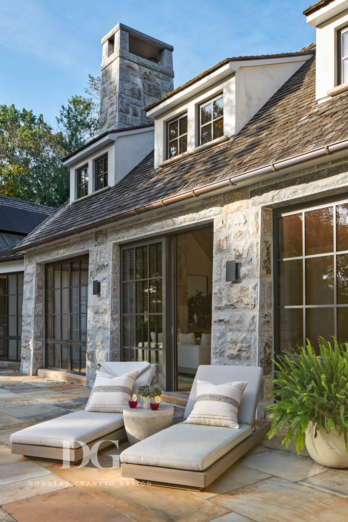 A patio with 2 wood loungers with neutral cushions and pillows. Side of a stone clad house with large black trimmed windows and sliding doors